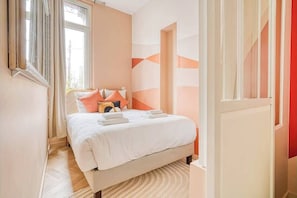 Ein ordentlich bezogenes Bett mit Kissen und Decke, ein Fenster mit Vorhängen und eine Wand mit abstrakten Mustern.