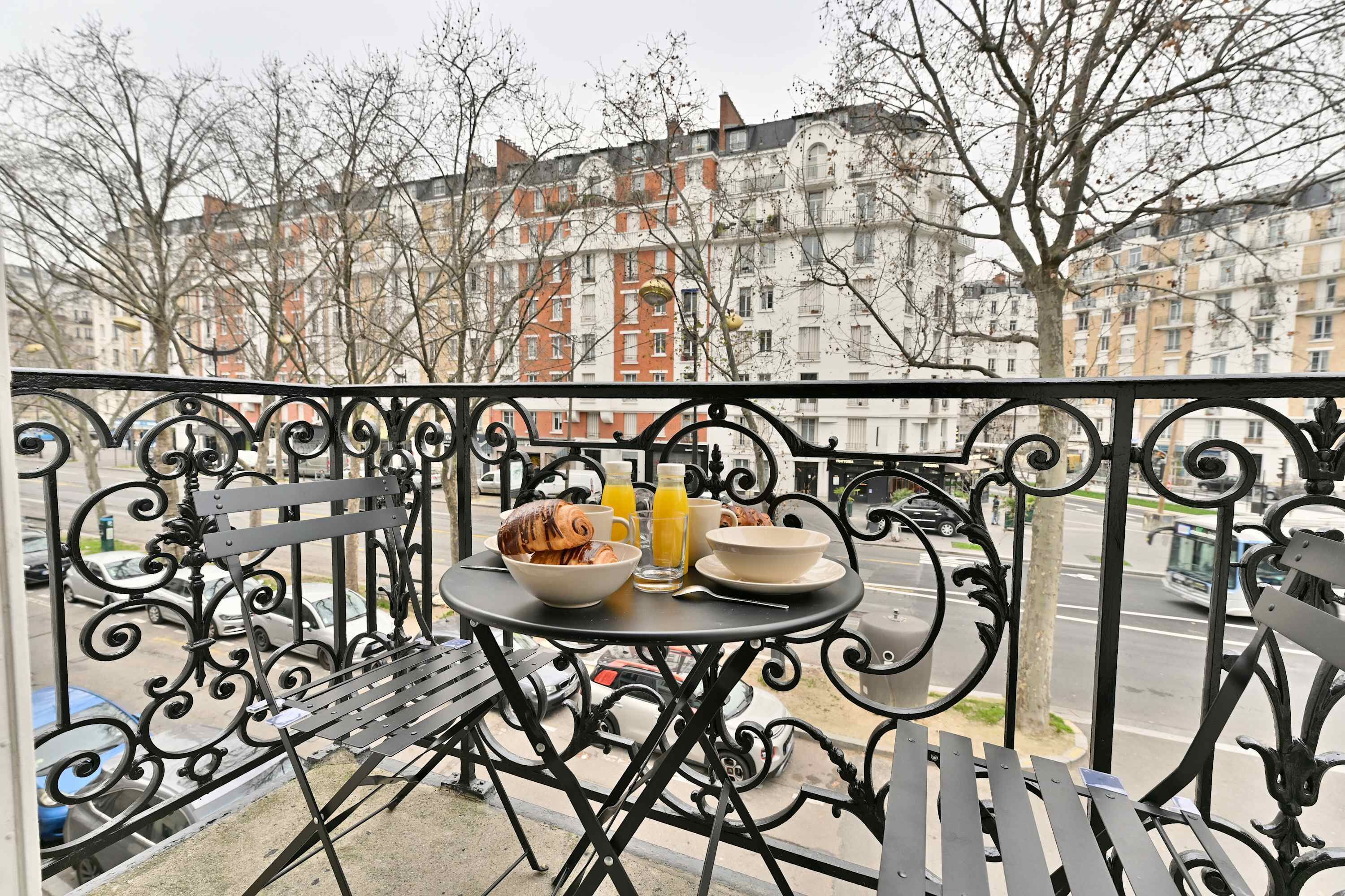 Ein Balkon mit einem Tisch, gedeckt für ein Frühstück, mit einem Croissant, einer Schüssel und zwei Gläsern Orangensaft.