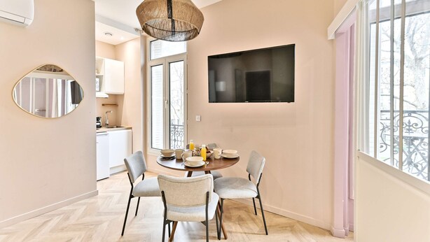 A modern dining area with a round table, two chairs, a television mounted on the wall, and a large window providing a view outside. - Charmant, confortable et climatisé - Paris 17 (Paris)