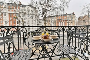 Ein Balkon mit Tisch, gedeckt für ein Frühstück, darunter eine Schüssel mit Müsli, eine Orangensaftflasche und ein Brotlaib.