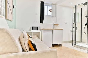 A modern, well-lit room with a sofa, a small kitchenette, a glass shower enclosure, and a window with a curtain. - Studio tout confort climatisé – proche métro (Paris)