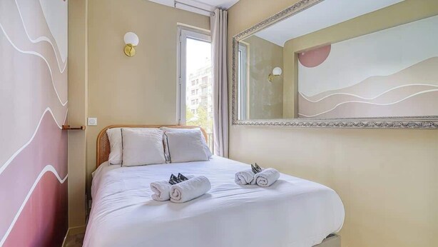 Uma cama bem arrumada com lençóis brancos e toalhas enroladas, um espelho grande com moldura ornamentada e uma parede com padrão de design ondulado.