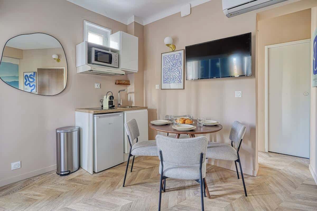 A compact kitchen with a microwave, a small refrigerator, and a sink. There is a dining table with four chairs and a TV mounted on the wall.