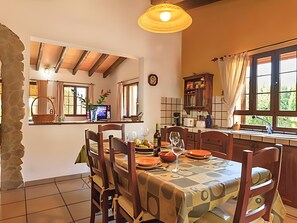 Un coin repas avec une table dressée pour quatre personnes, des chaises en bois, un lustre et une vue sur la cuisine par une fenêtre.