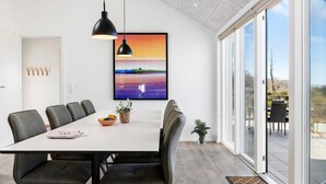 Un espace repas moderne avec une table blanche, des chaises noires, des luminaires suspendus, un tableau encadré et une vue dégagée sur l’extérieur grâce à de grandes portes vitrées.
