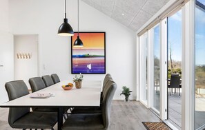 Zona pranzo moderna con tavolo bianco, sedie nere, illuminazione a sospensione, un quadro incorniciato e vista panoramica tramite ampie porte in vetro.