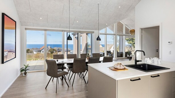 Une cuisine moderne dotée d’un grand îlot central, d’un coin repas avec des chaises et de grandes fenêtres offrant une vue sur la mer.