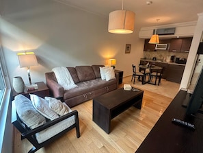 Ein modernes Wohnzimmer mit Sofa, Couchtisch und Sesseln, ein Küchenbereich mit Esstisch sowie ein großes Fenster, das für natürliches Licht sorgt.