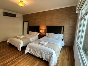 Ein Hotelzimmer mit zwei Betten, Holzboden, einem großen Fenster und einer Wand-Klimaanlage.