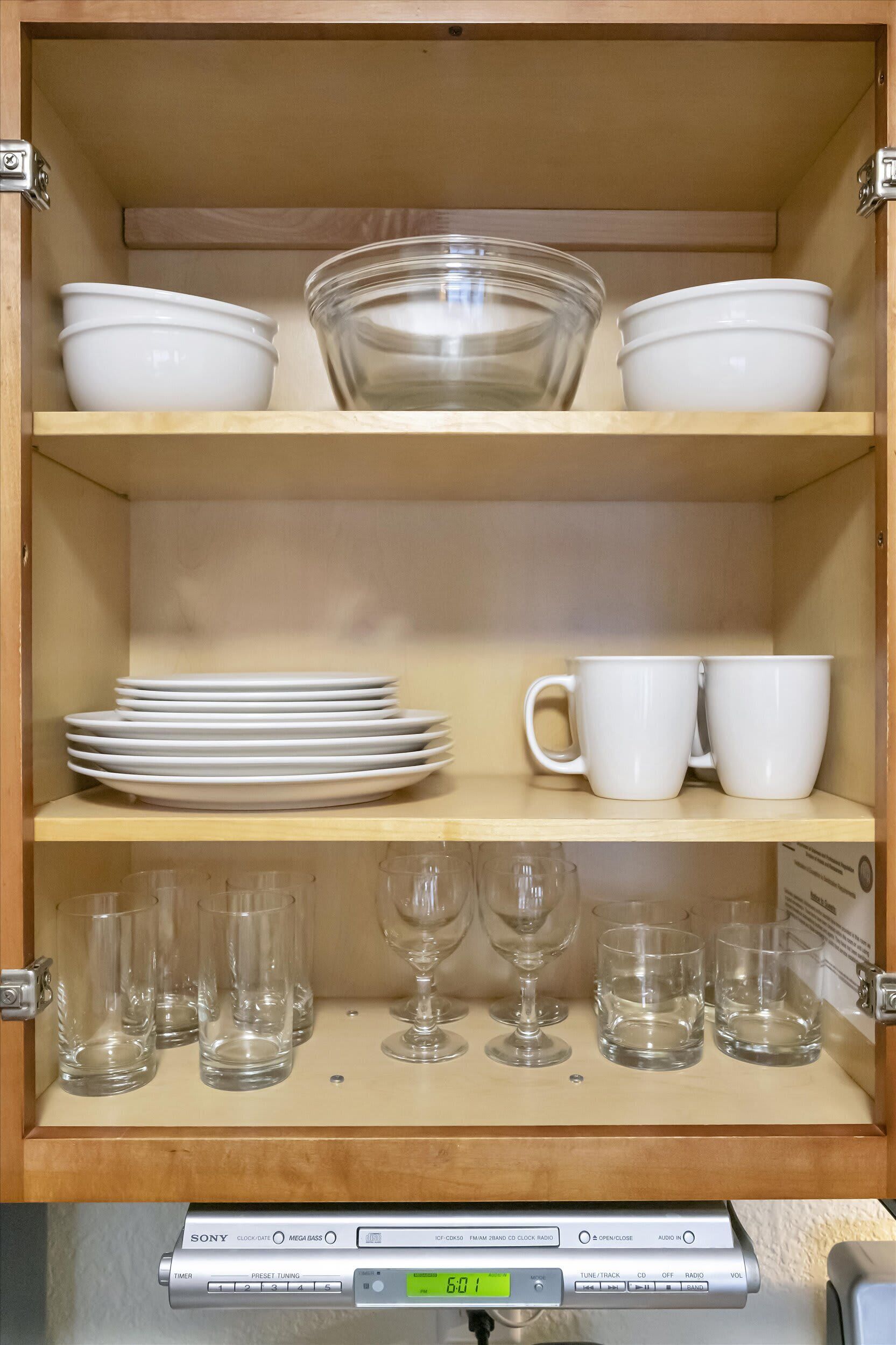 Un meuble de cuisine avec des étagères où sont rangés des bols, des assiettes et des tasses blancs. En dessous, on trouve des verres à vin et une horloge Sony.