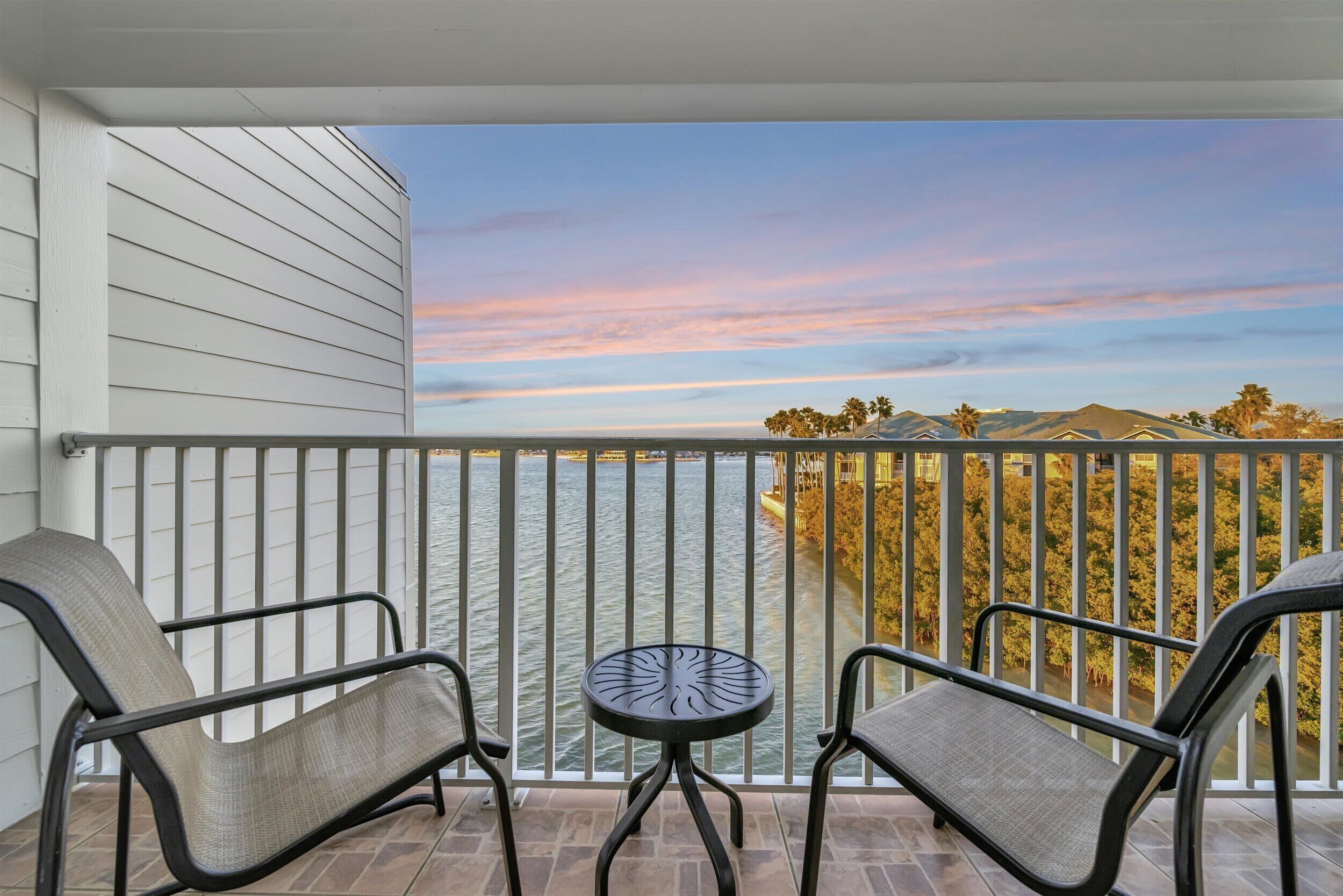 A balcony with two chairs and a small table, overlooking a body of water and a distant landscape with trees and hills.