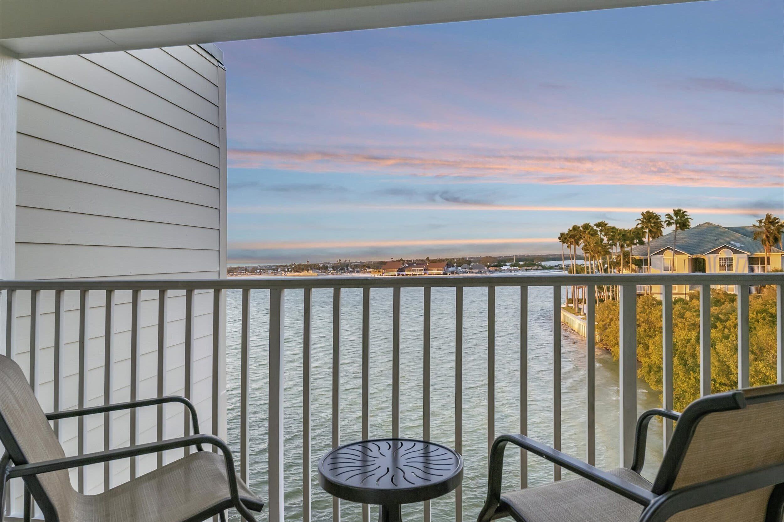 A balcony with two chairs and a small table, overlooking a body of water with a distant shoreline and palm trees.
