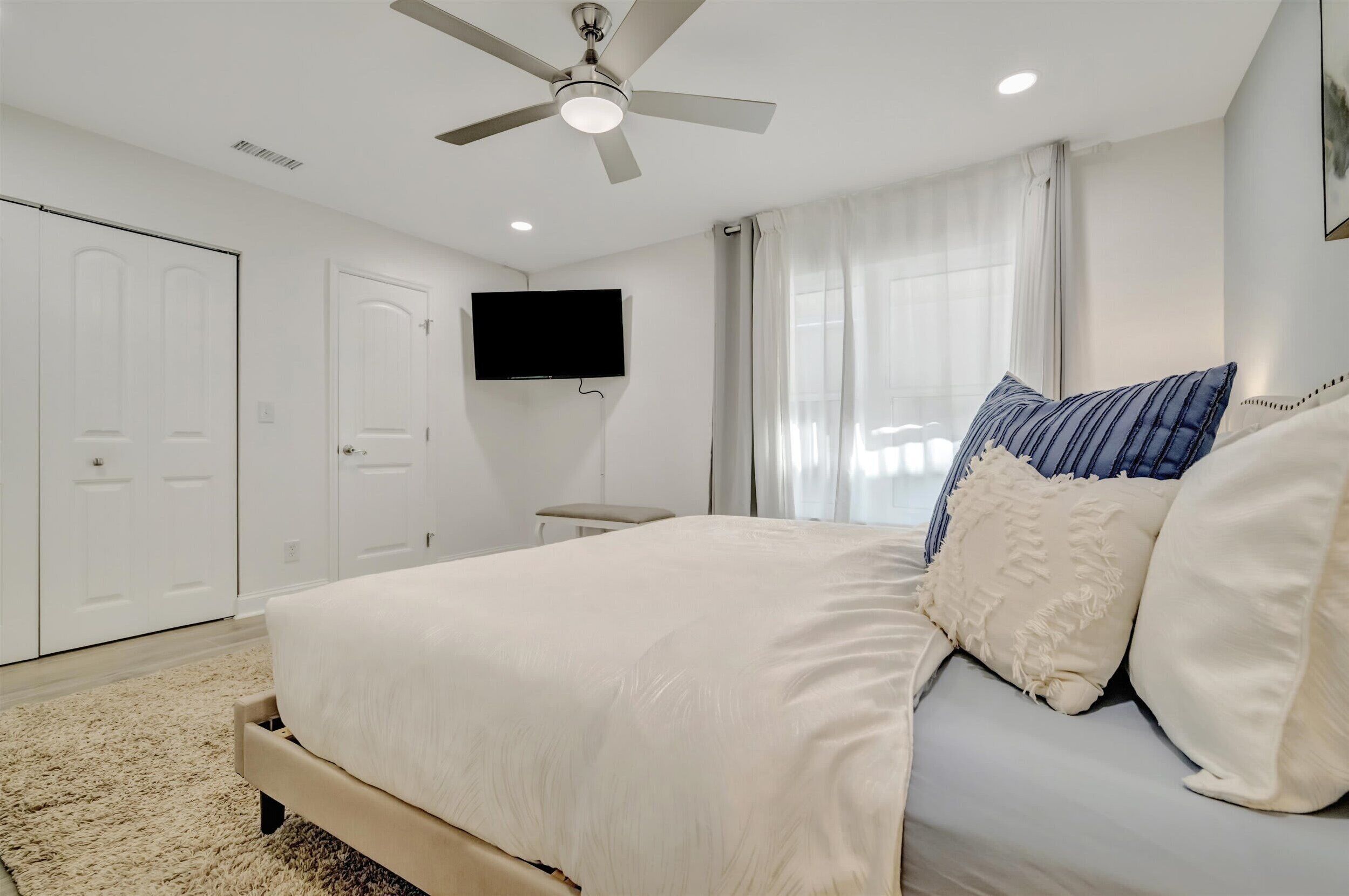 Un dormitorio con cama, ventilador de techo, televisión y ventana con cortinas.