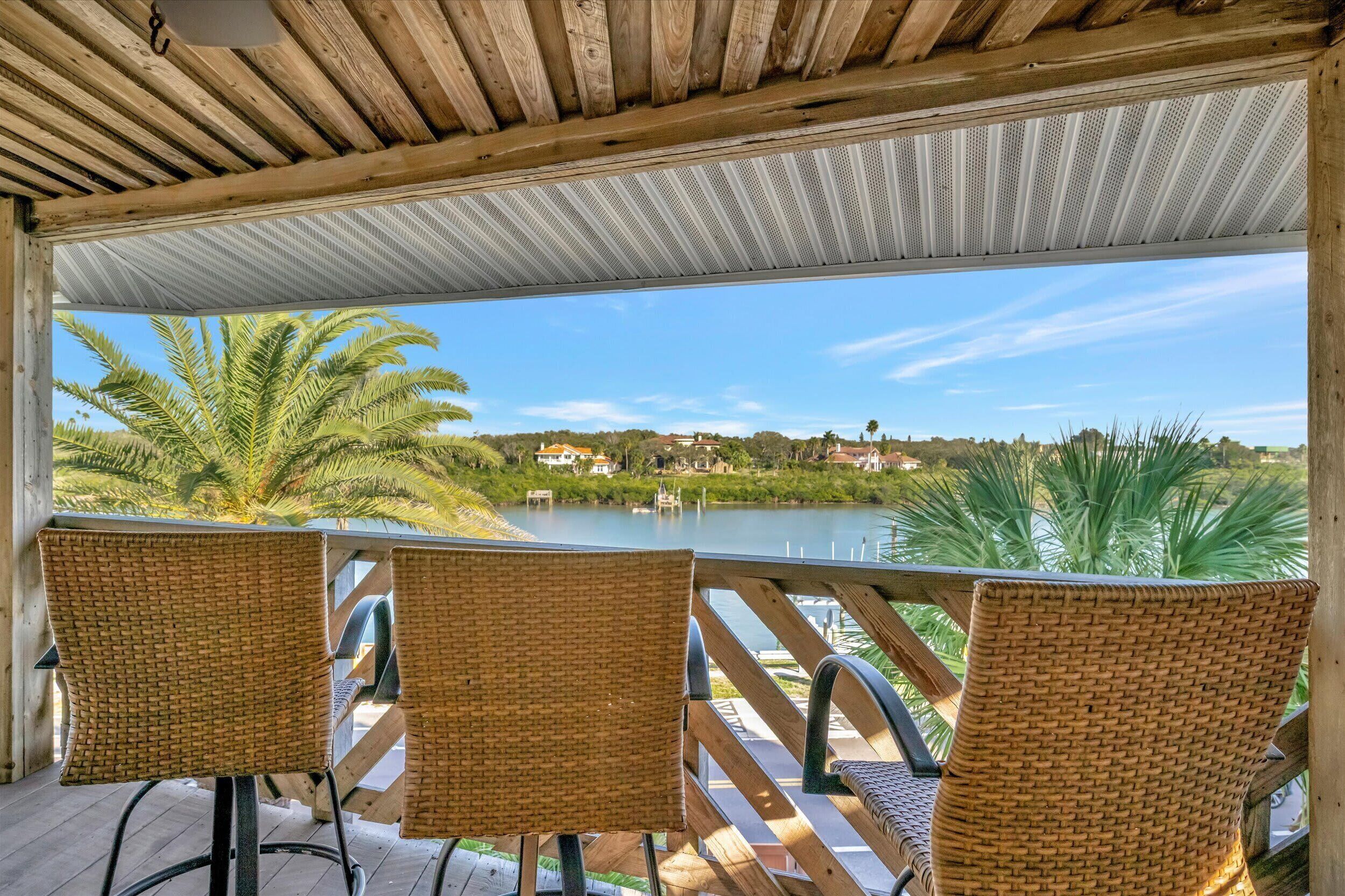 A covered outdoor seating area with wicker chairs and a view of a lake and distant houses.