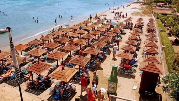 A beach with rows of wooden umbrellas and lounge chairs, a clear view of the sea, and mountains in the background. - Villa Amorata - Schinias Private Garden Retreat (Marathon)