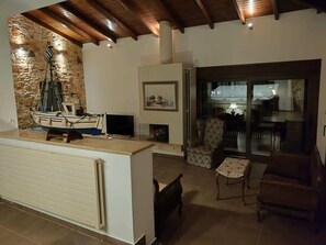 A living room with a stone wall, a fireplace, a television, and a dining area with a glass door leading outside. - Villa Amorata - Schinias Private Garden Retreat (Marathon)