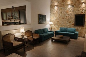 A cozy living room with a stone wall, a wooden ship model, a rattan chair, two teal sofas, and a coffee table. - Villa Amorata - Schinias Private Garden Retreat (Marathon)
