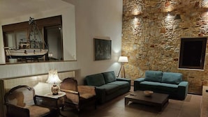 A living room with a stone wall, two green sofas, a wooden chair, a small table with a lamp, and a ship model on the wall. - Villa Amorata - Schinias Private Garden Retreat (Marathon)