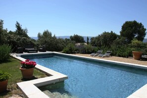 A rectangular swimming pool with a clear blue water surface, surrounded by a low stone wall. There are two lounge chairs on the right side of the pool, and a potted plant with pink flowers is visible on the left. - La Villa des Restanques (Cabrieres-d'Avignon)