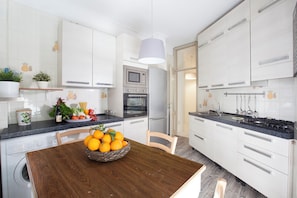A modern kitchen with white cabinets, a wooden dining table, and a bowl of oranges. - YourHome - Parco Sereno Apartment (Sorrento)