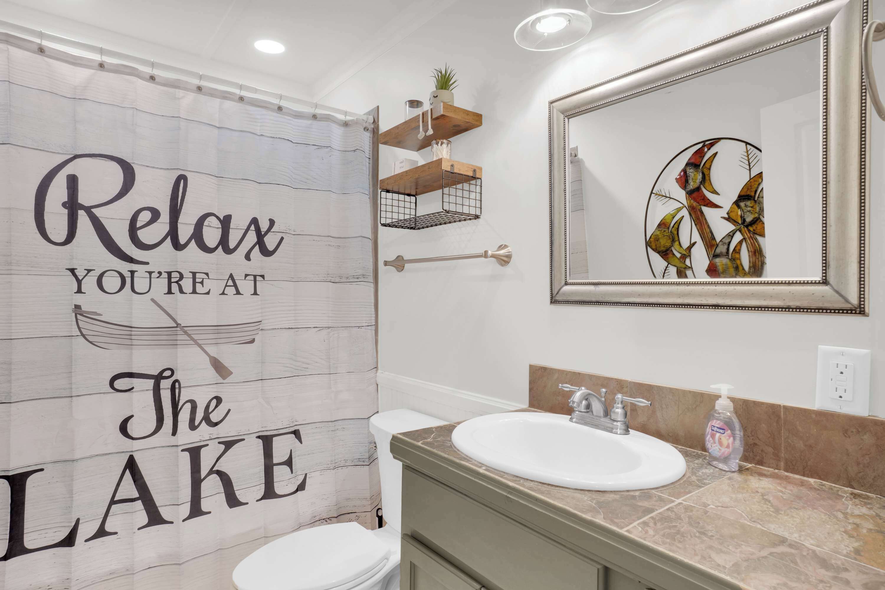 Een badkamer met een douchegordijn waarop staat 'Relax, je bent aan het meer', een wastafel met spiegel erboven en een decoratief wandobject.