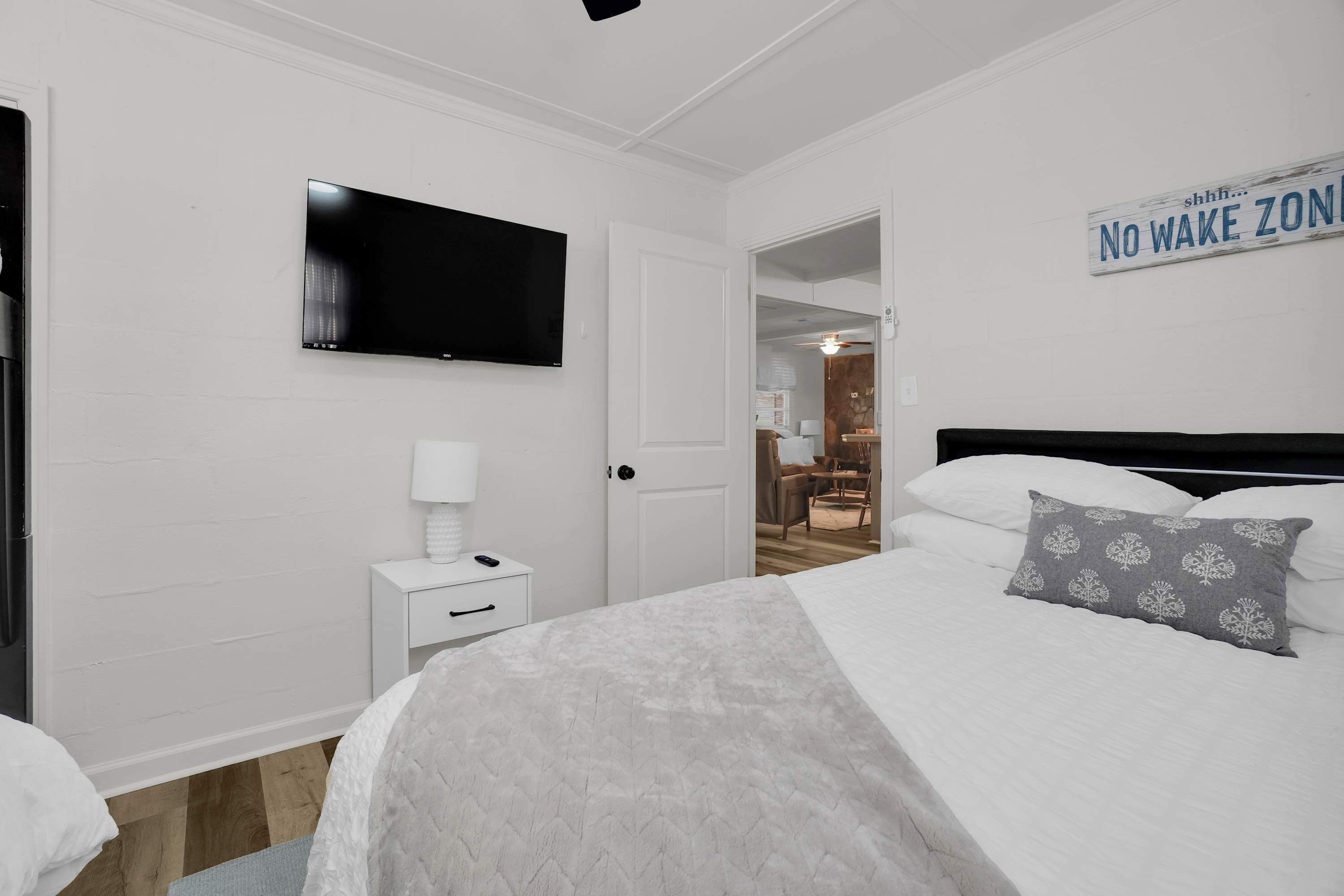 A bedroom with a bed, a TV mounted on the wall, a nightstand with a lamp, and a door leading to another room.