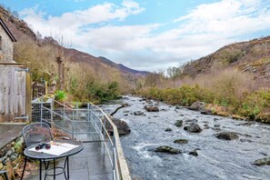 Un patio junto al río con una mesa, sillas y barandilla con vista a un río que fluye.