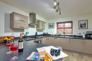 Une cuisine moderne équipée d’un four encastré, d’un évier et d’une fenêtre donnant sur un beau paysage. On peut voir deux verres à vin sur le comptoir, une bouteille de vin et une carte.