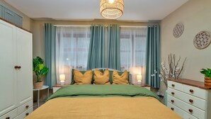 Quarto com cama, roupa de cama verde e amarela, mesa de cabeceira com abajur, cômoda e janela com cortinas.