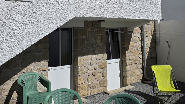 Un edificio de dos pisos con balcón, paredes de piedra, y una mesa con sillas y una jarrón.