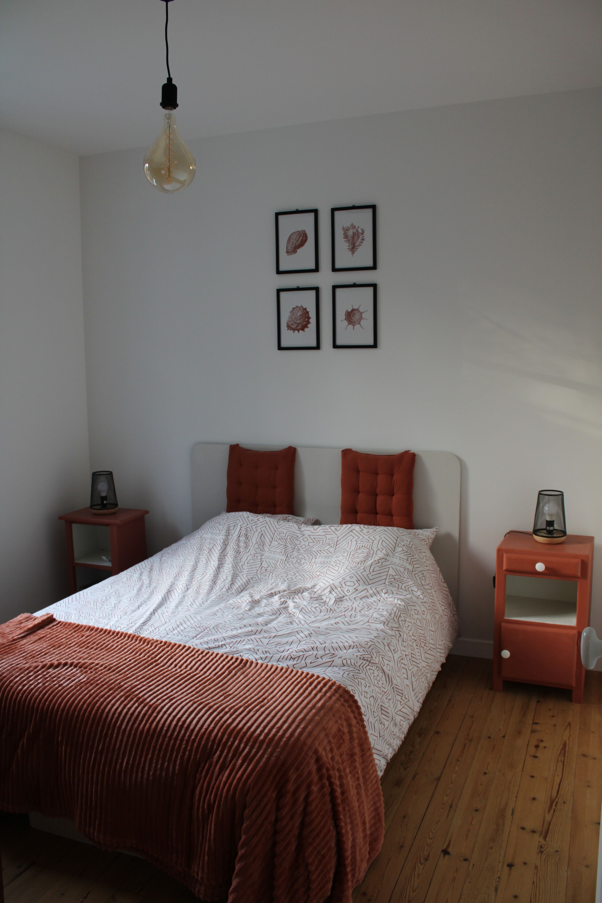 Um quarto com uma cama, duas mesas de cabeceira, uma luminária pendente e obras de arte emolduradas na parede.