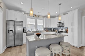 A modern kitchen with white cabinets, a stainless steel refrigerator, a built-in oven, a microwave, and a large island with stools. - Beautiful 5 Bedroom oceanfront home with community pool (North Topsail Beach)