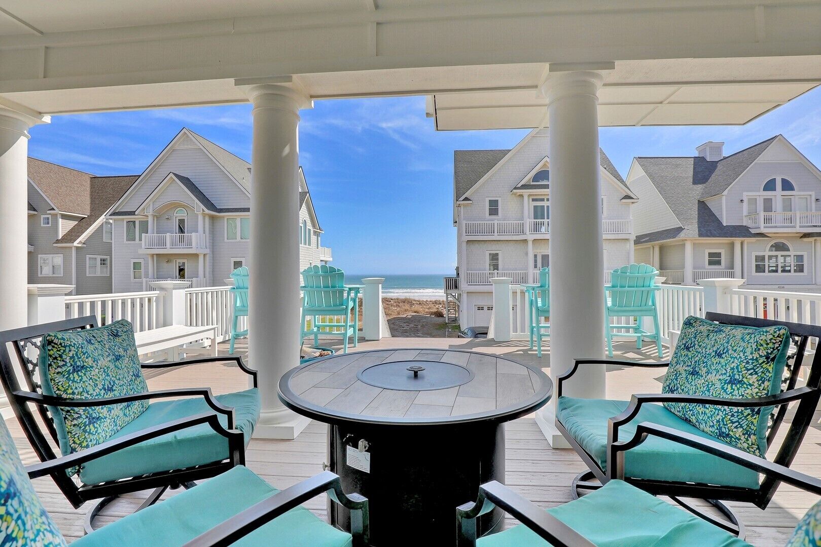 A covered outdoor seating area with a round table and chairs, overlooking a beach with multiple buildings.