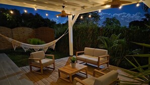 Ett trädäck med utemöbler, inklusive en soffa och stolar, under en pergola med ljusslingor.