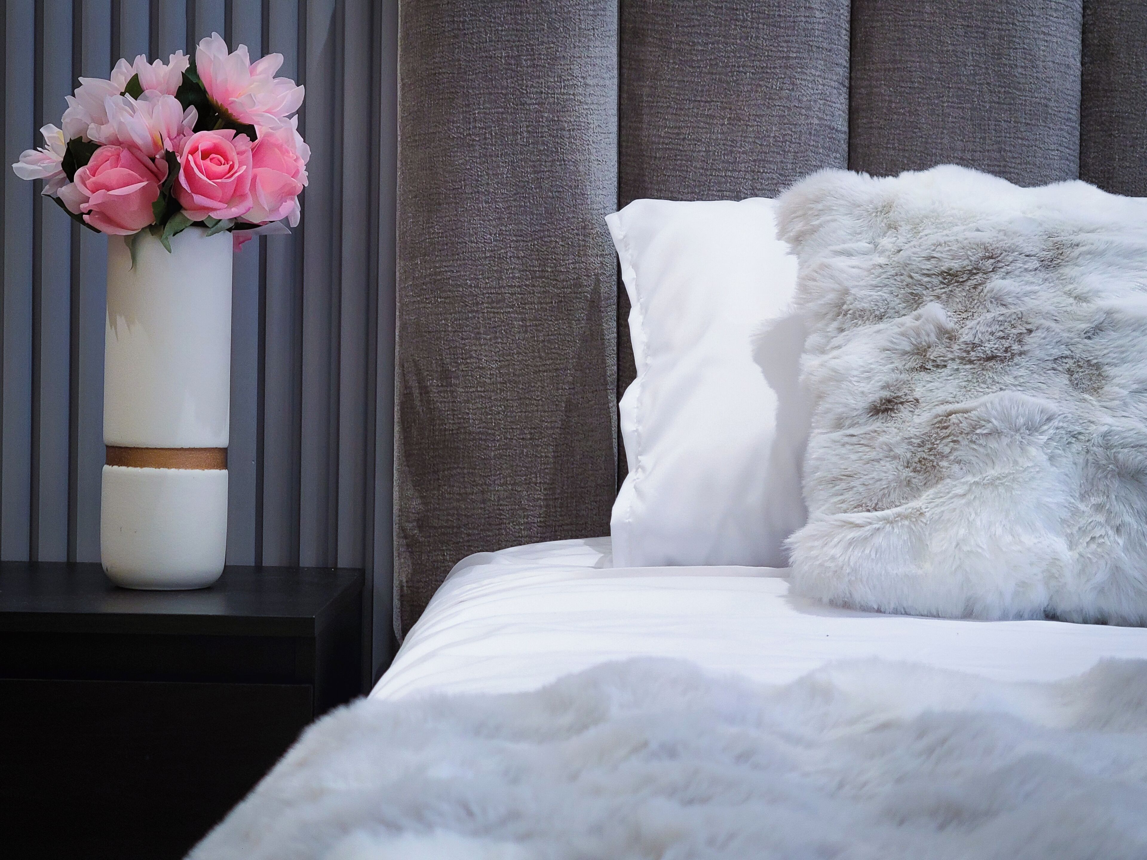 A neatly made bed with white bedding and a fluffy pillow, a white vase with pink flowers on a bedside table, and a grey upholstered headboard.