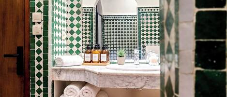 A bathroom with a green and white checkered tile backsplash, a marble countertop, and a mirror above it. There are towels, a small plant, and toiletries on the counter.