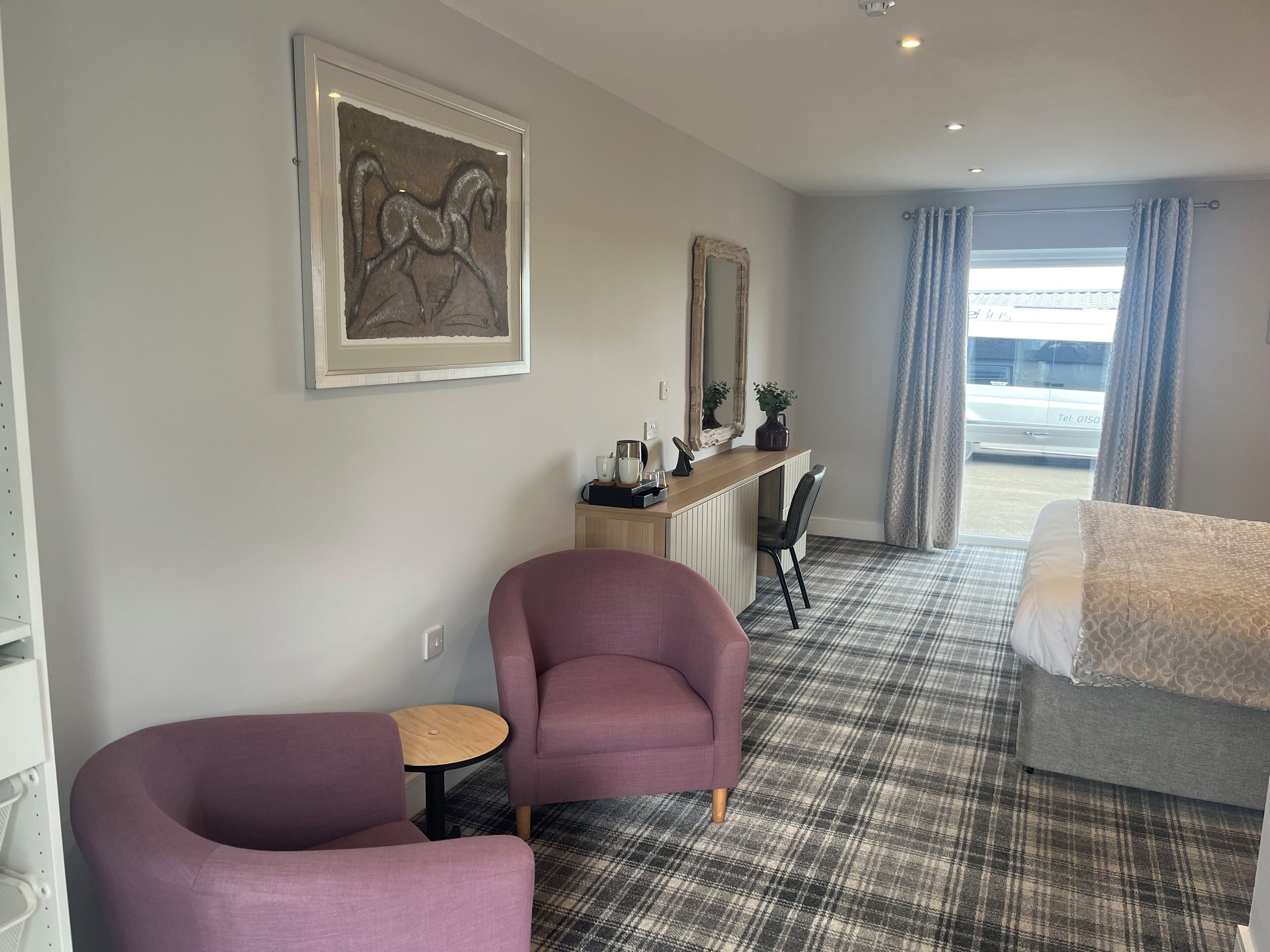 Une chambre d’hôtel comprenant un lit, deux fauteuils, une petite table, un bureau avec un miroir, une machine à café et une œuvre d’art encadrée accrochée au mur.