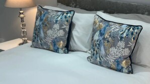 Una cama con dos almohadones decorativos de estampado floral azul y dorado, una lámpara de noche y un cabecero gris.