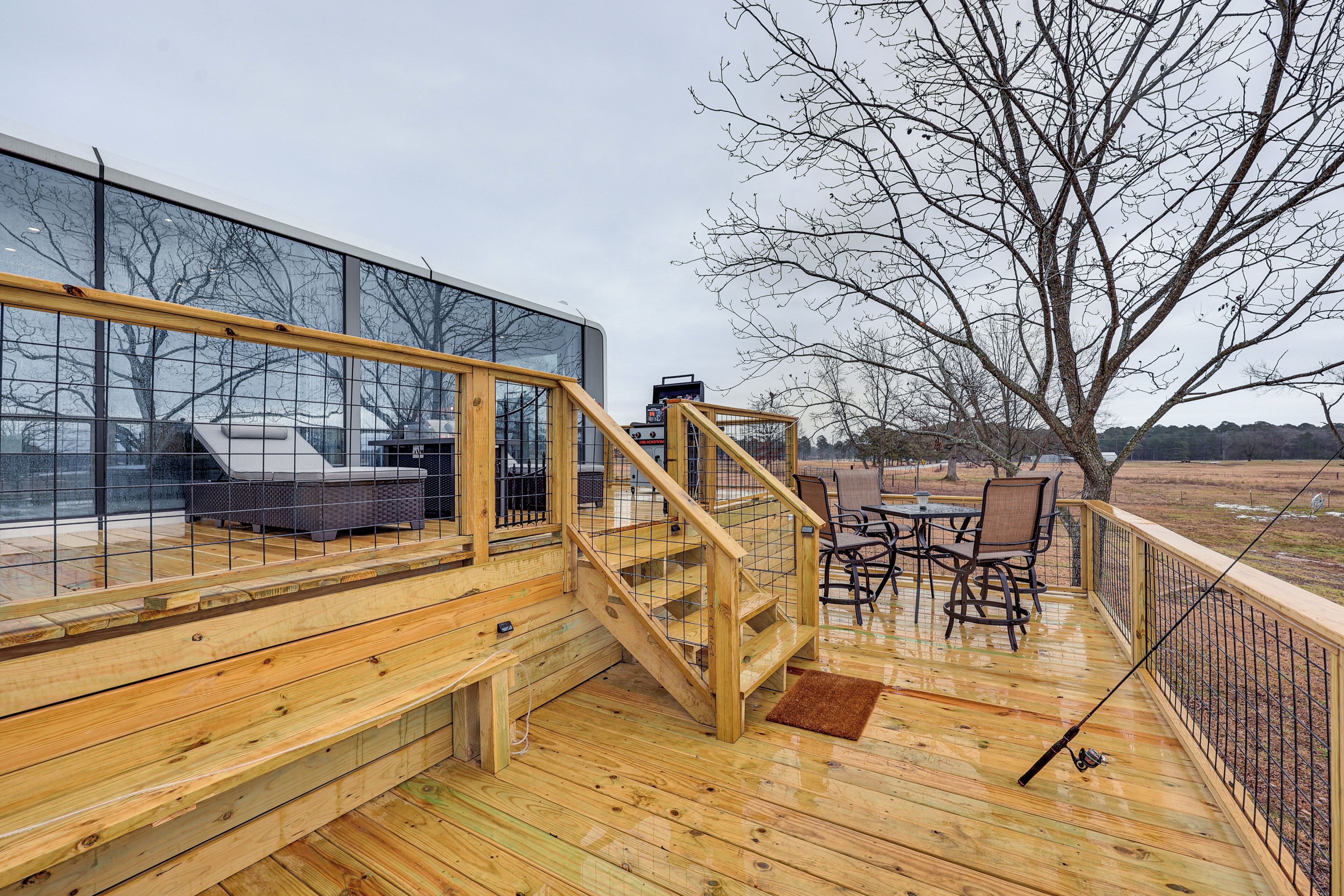 Una terraza de madera con una escalera que lleva a un área con paredes de vidrio, mobiliario exterior y vistas del paisaje circundante.