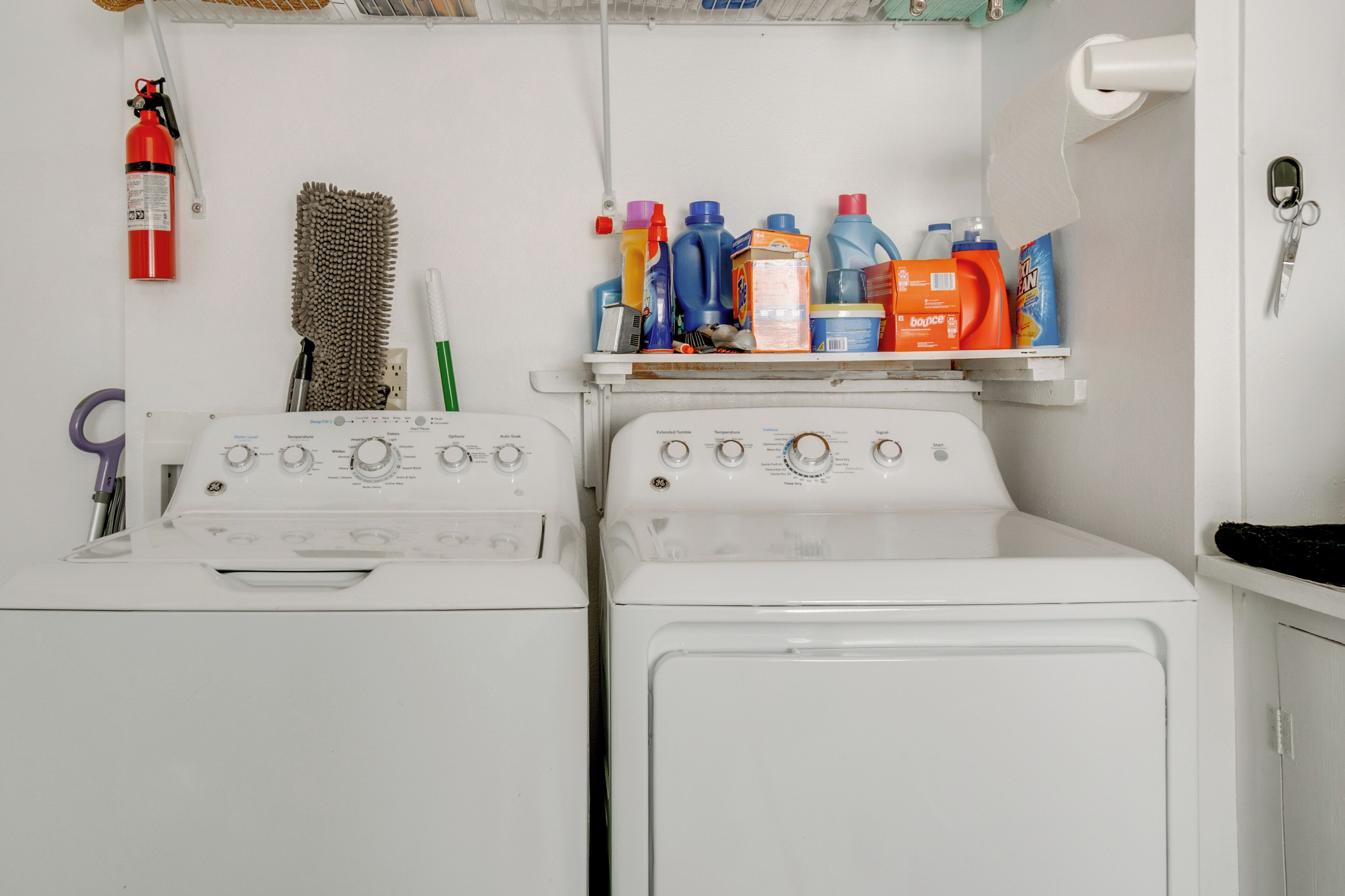 A laundry area with two white washing machines, a shelf with various cleaning supplies, a fire extinguisher, and a hanging brush.