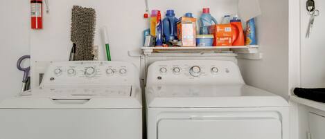 A laundry area with two white washing machines, a shelf with various cleaning supplies, a fire extinguisher, and a hanging brush.
