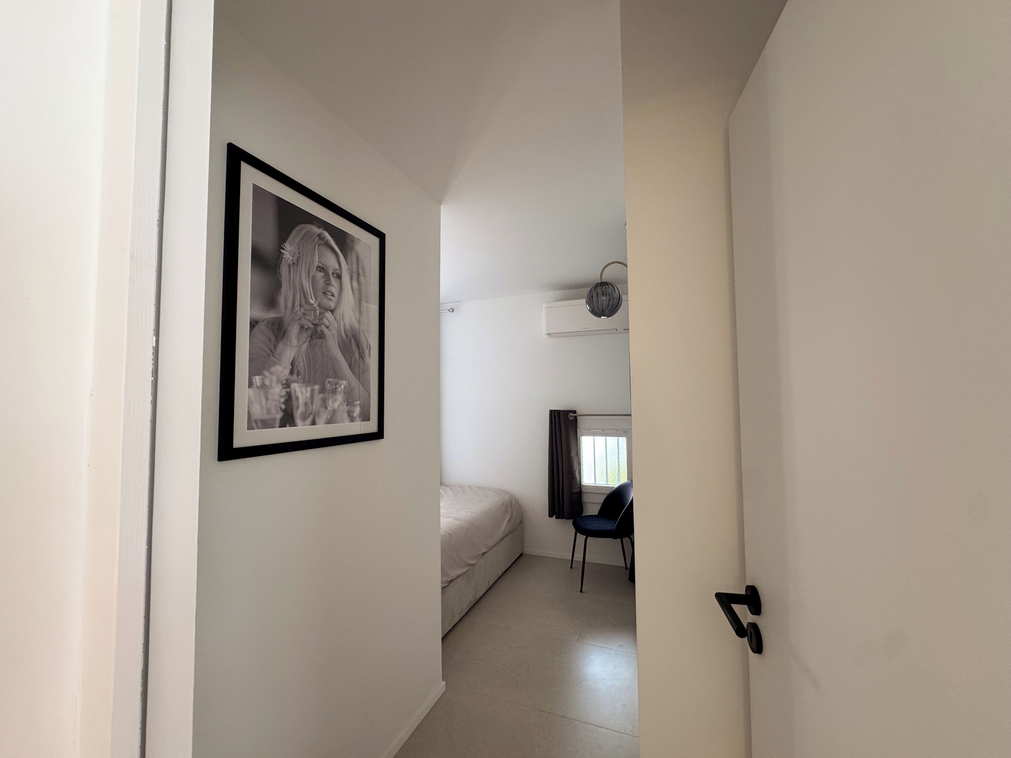 Un couloir étroit, avec une photographie en noir et blanc encadrée sur le mur de gauche, un lit avec un matelas blanc et une chaise sombre au fond, et une porte avec une poignée noire sur le côté droit.