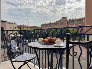 Un balcón con una mesa dispuesta para dos personas, con pasteles y una taza, con vistas a una calle con edificios.