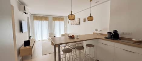 A modern kitchen with a central island, bar stools, a dining area, and a living space with a sofa and curtains.