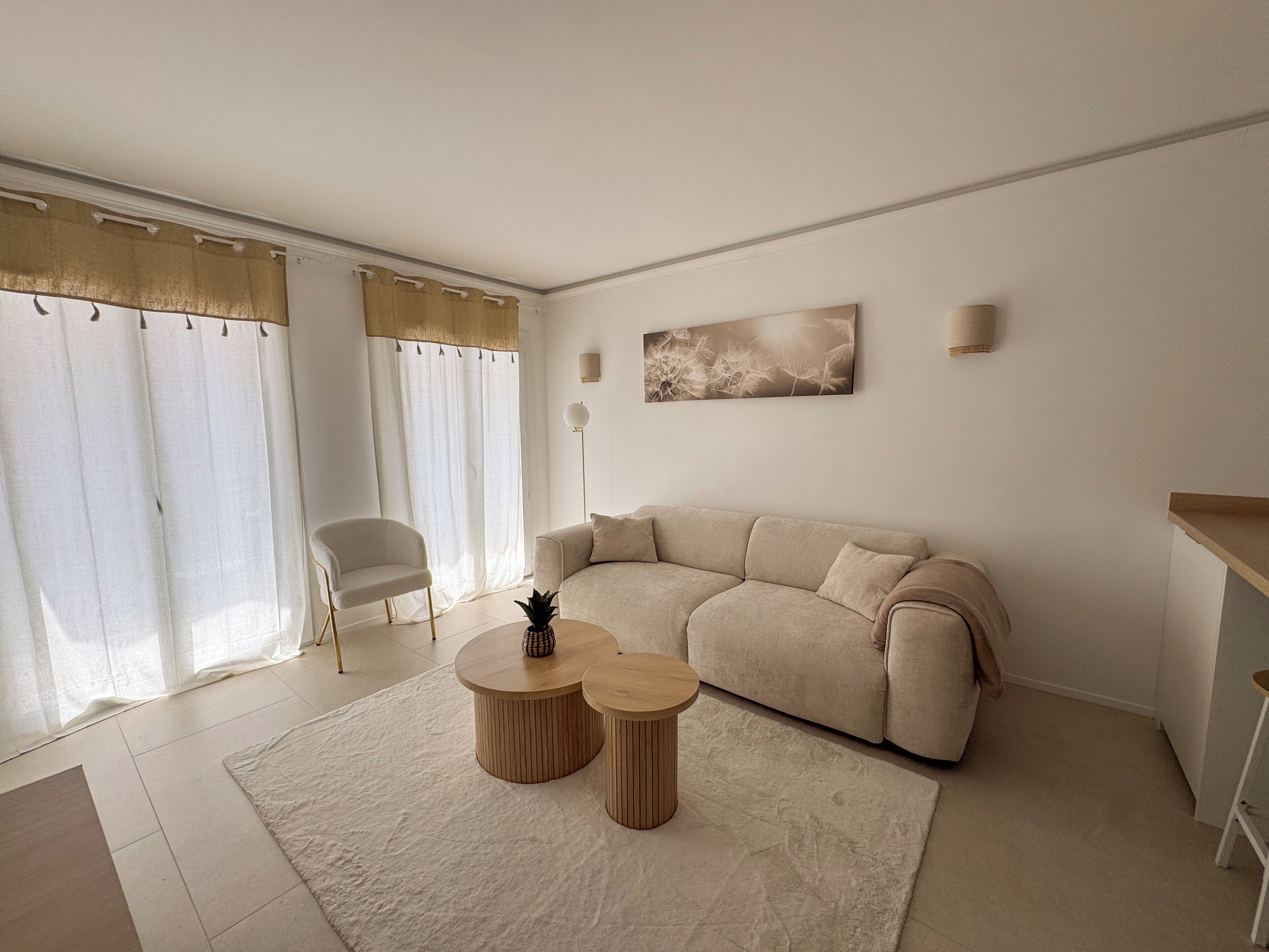 Un salón moderno con un sofá color beige, dos mesitas redondas de madera, una planta en maceta y un amplio ventanal con cortinas de gasa.