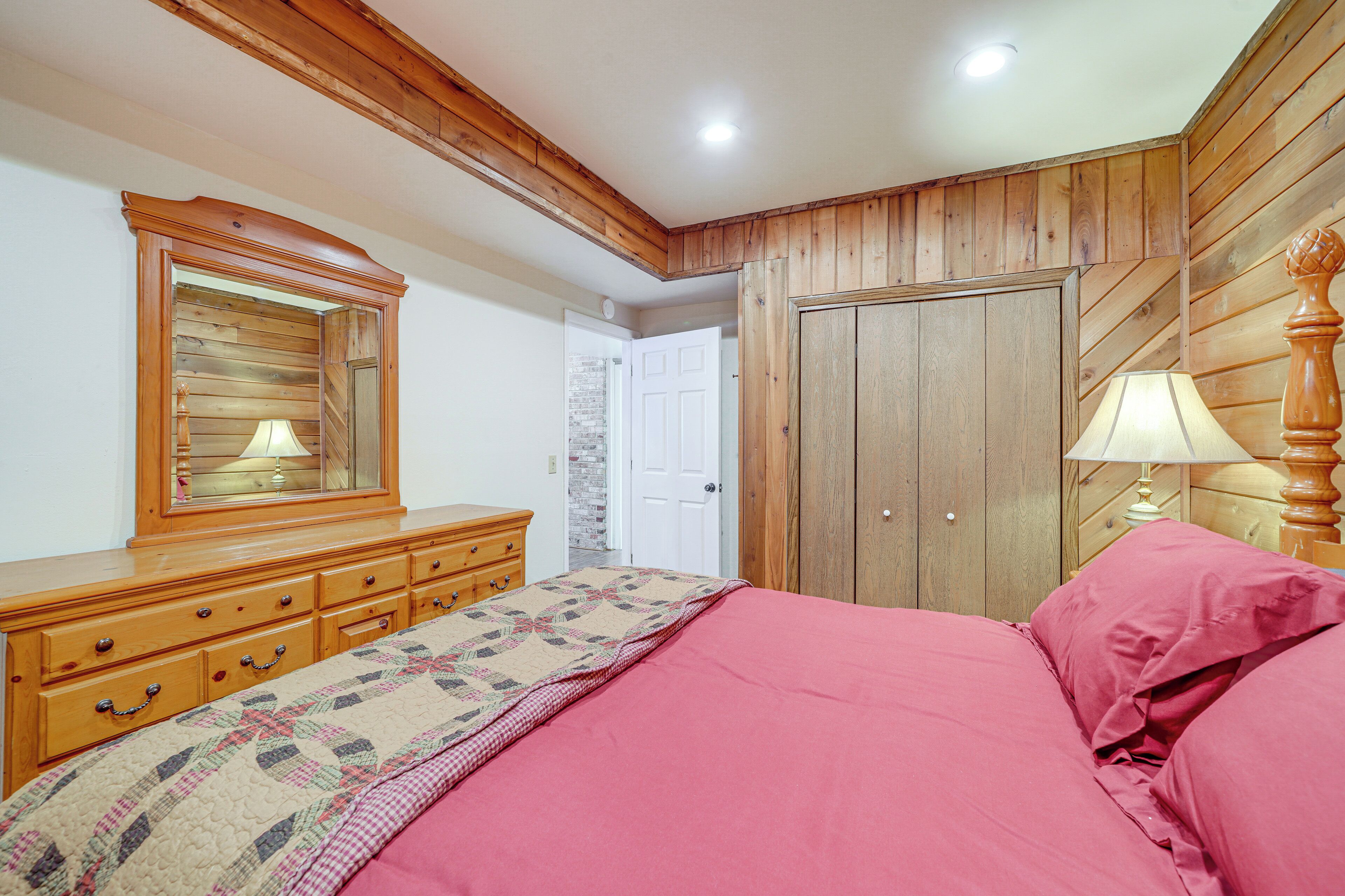 A bedroom with wooden paneling, a bed with pink bedding, a dresser with drawers, a mirror, and a door leading to another room.