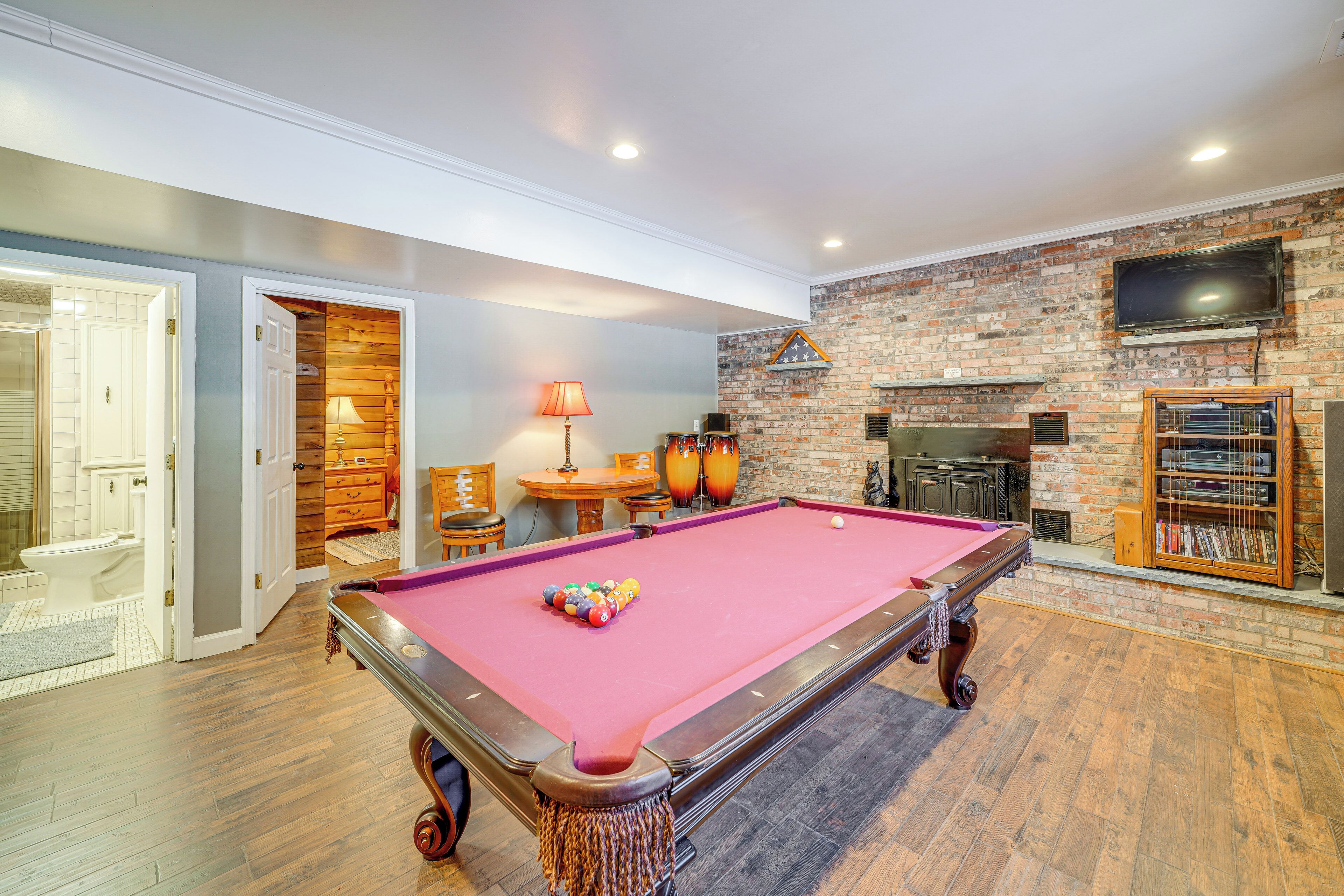 A pool table in a room with a brick wall, a television mounted above it, and a fireplace.