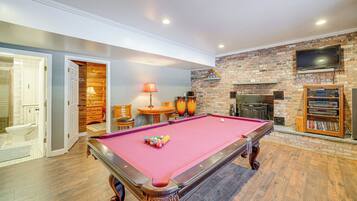 A pool table in a room with a brick wall, a television mounted above it, and a fireplace.