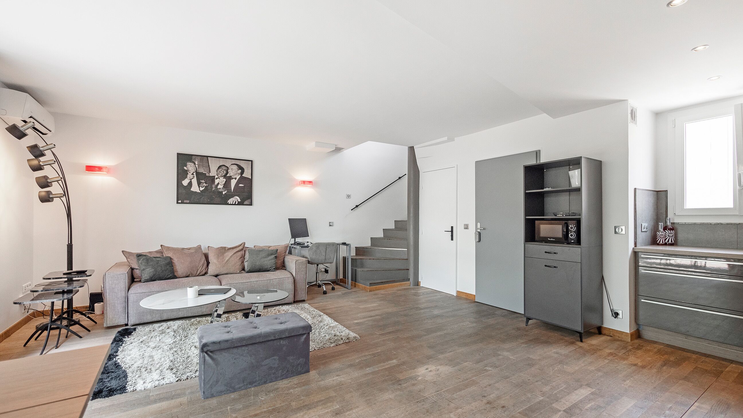 A modern living room with a grey sofa, a coffee table, and a grey ottoman. There is a staircase leading to another level, a grey cabinet with a microwave, and a framed picture on the wall.
