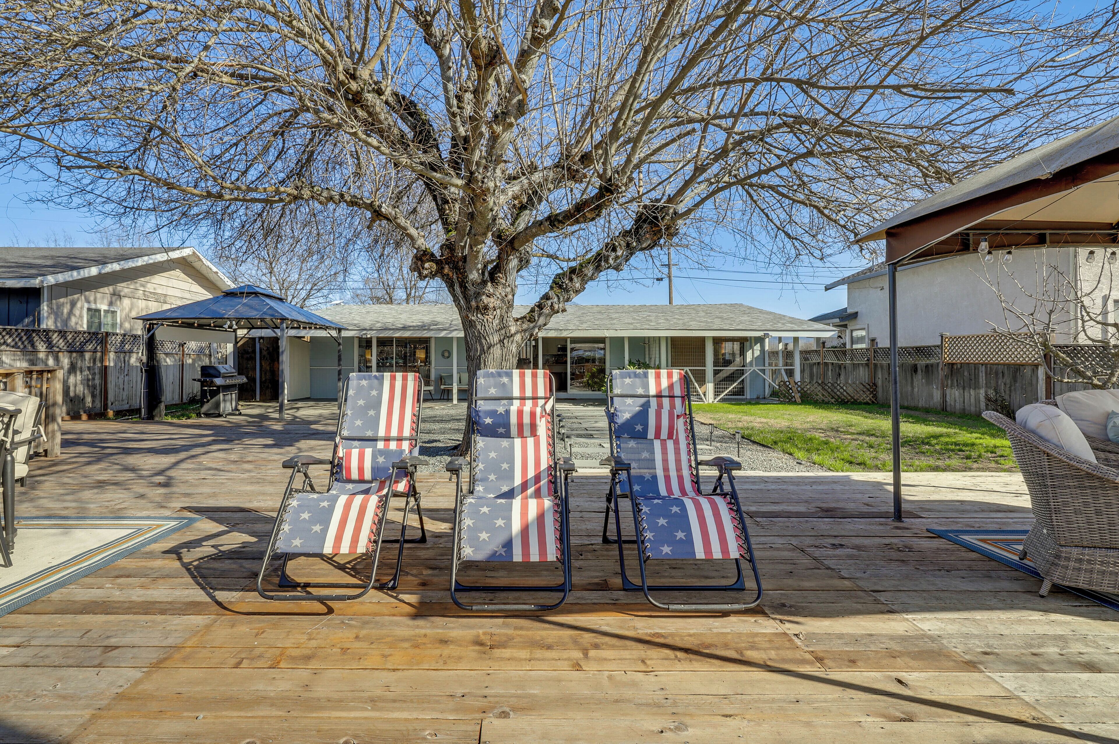 A backyard with three lounge chairs featuring an American flag pattern, a large tree, and a covered patio area.